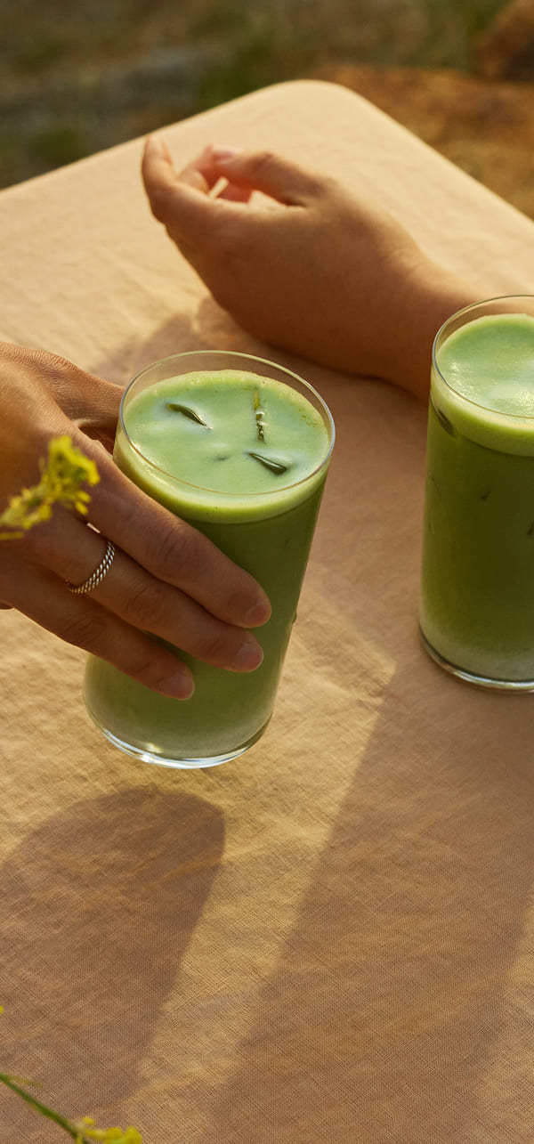 Two glasses of green iced matcha drink on a beige tablecloth with a person's hands visible holding one glass, in warm natural light.