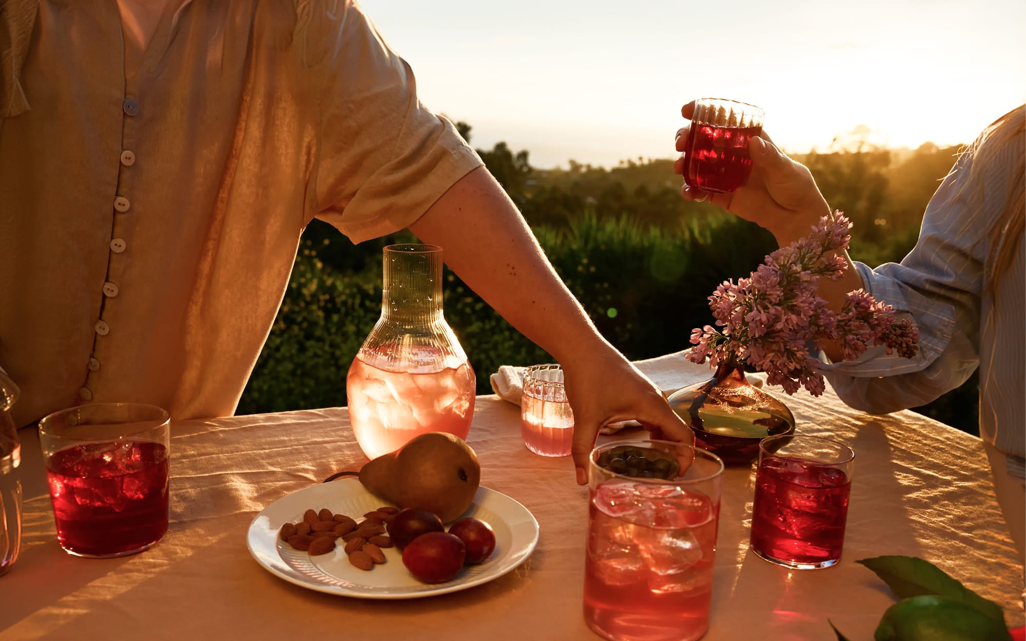 Two people at a table outdoors, enjoying a sunset with drinks and snacks.