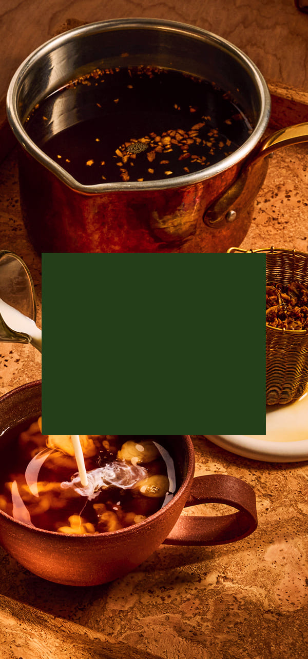 A warm, rustic scene showing a dark liquid, likely coffee or tea, in a copper pot and a matching copper mug with milk being poured in. A dark green square overlay obscures the center of the composition.