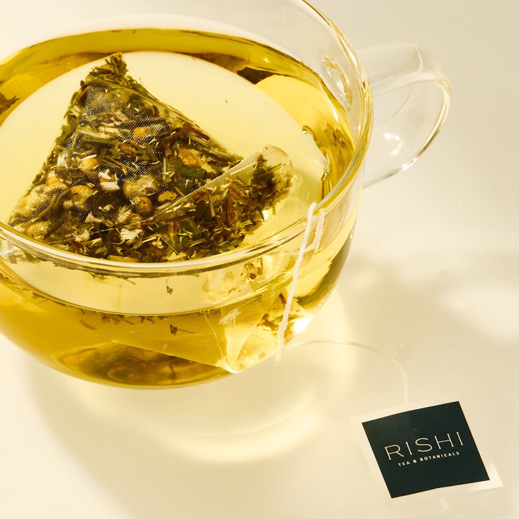 A glass cup of herbal tea brewed with a pyramid-shaped organic tea sachet from Rishi Tea & Botanicals, the tag resting on the table, showcases their premium ingredients.