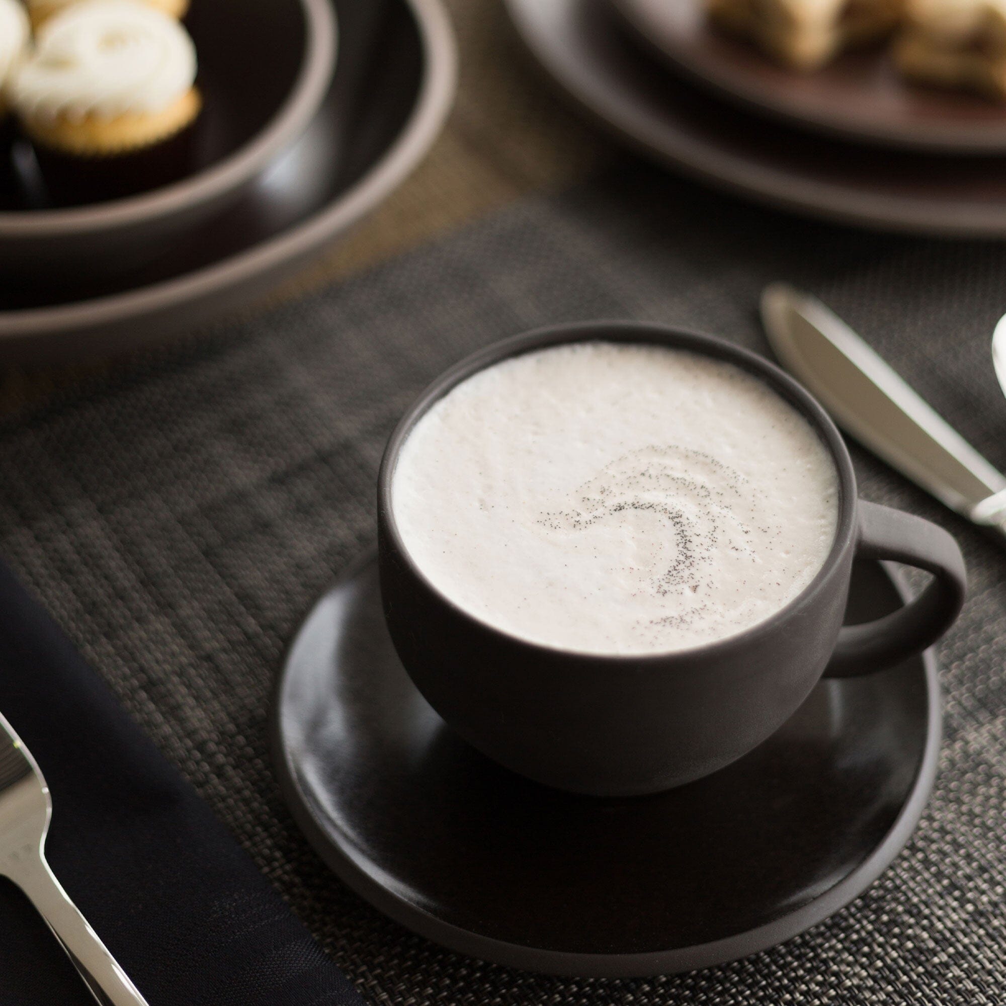 A cup of cappuccino with frothy milk on top sprinkled with Rishi Tea & Botanicals Sweet Houjicha Cocoa Powder, served on a dark saucer, with desserts in the background.