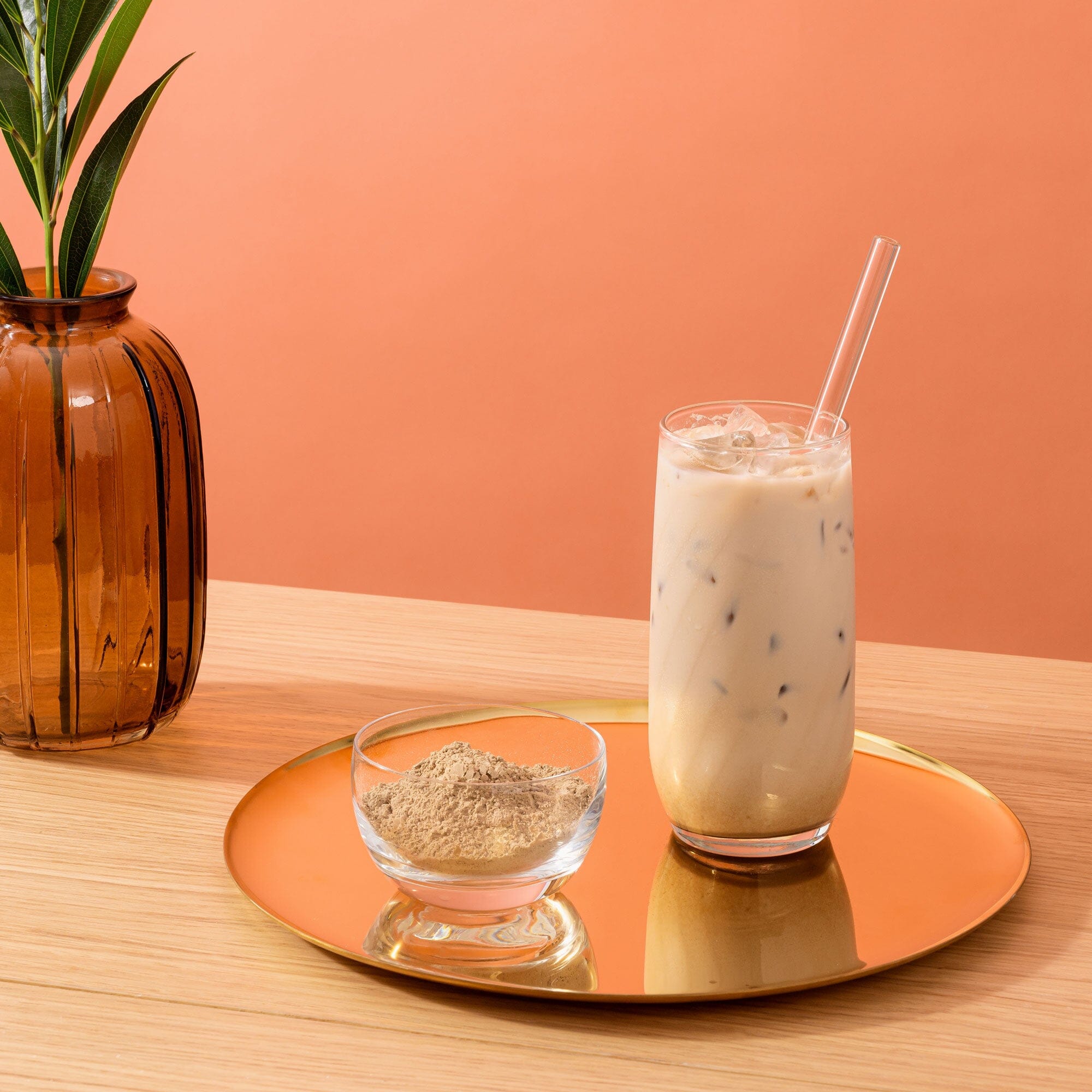 A glass of iced coffee with a straw, accompanied by a bowl of Sweet Houjicha Cocoa Powder, placed on a reflective orange surface with a plant in the background from Rishi Tea & Botanicals.