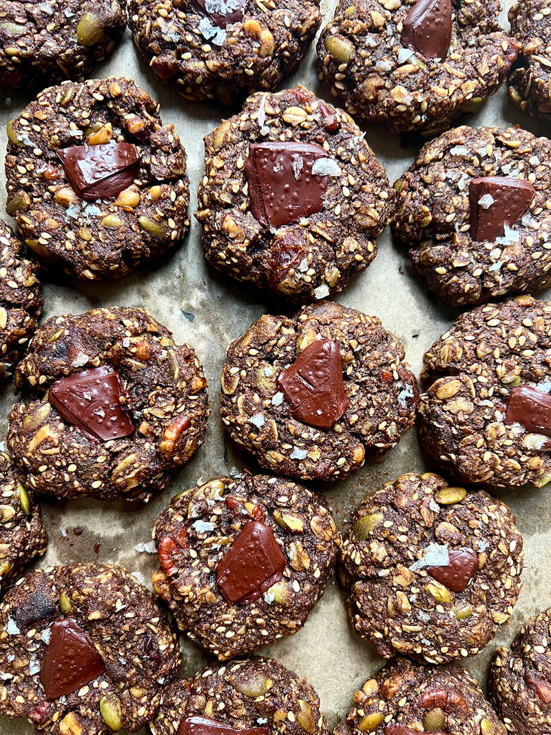 A top-down view of multiple cookies arranged closely together on parchment paper. Each cookie is studded with oats, seeds, and flakes, and topped with a chunk of dark chocolate.
