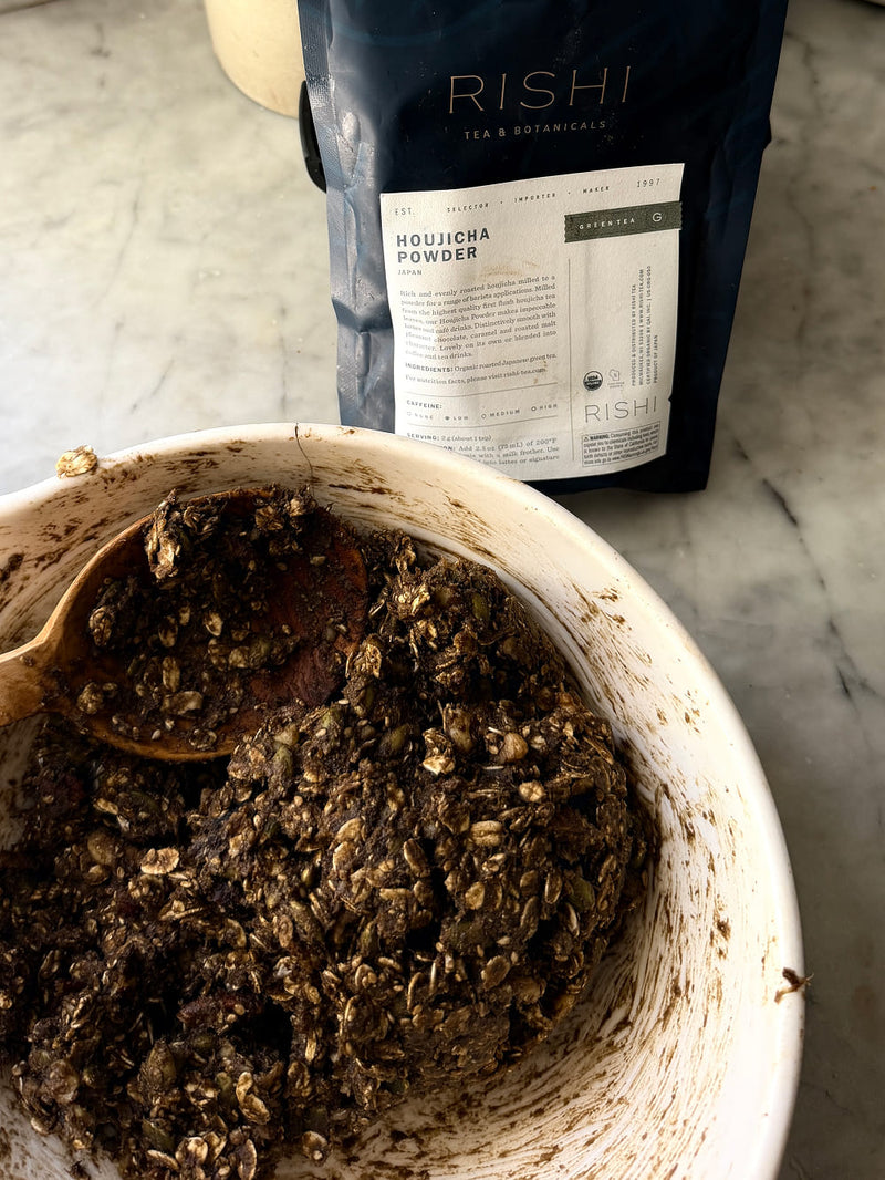 A bag of Rishi Houjicha Powder tea stands behind a white bowl filled with a dark brown seeds-tea mixture on a light marble countertop.
