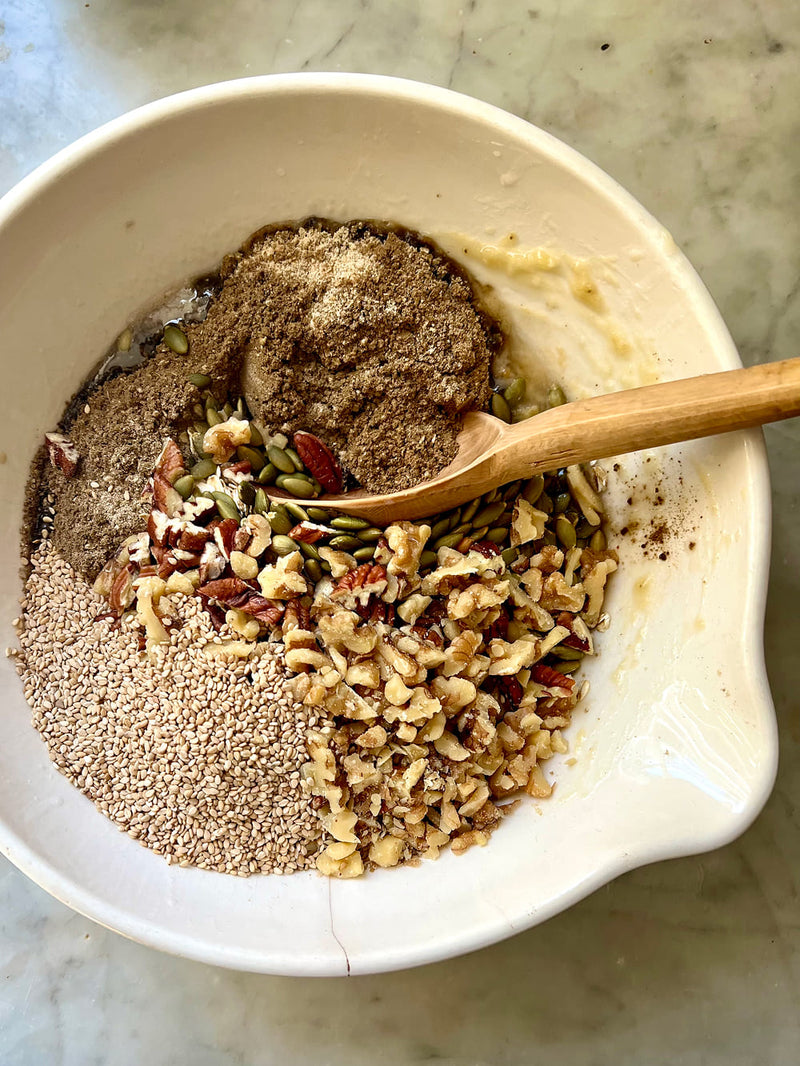 A white ceramic bowl containing mixed baking ingredients, including chopped walnuts, pumpkin seeds, chia seeds, and a brown powder, with a wooden spoon resting inside, set on a light marble surface.