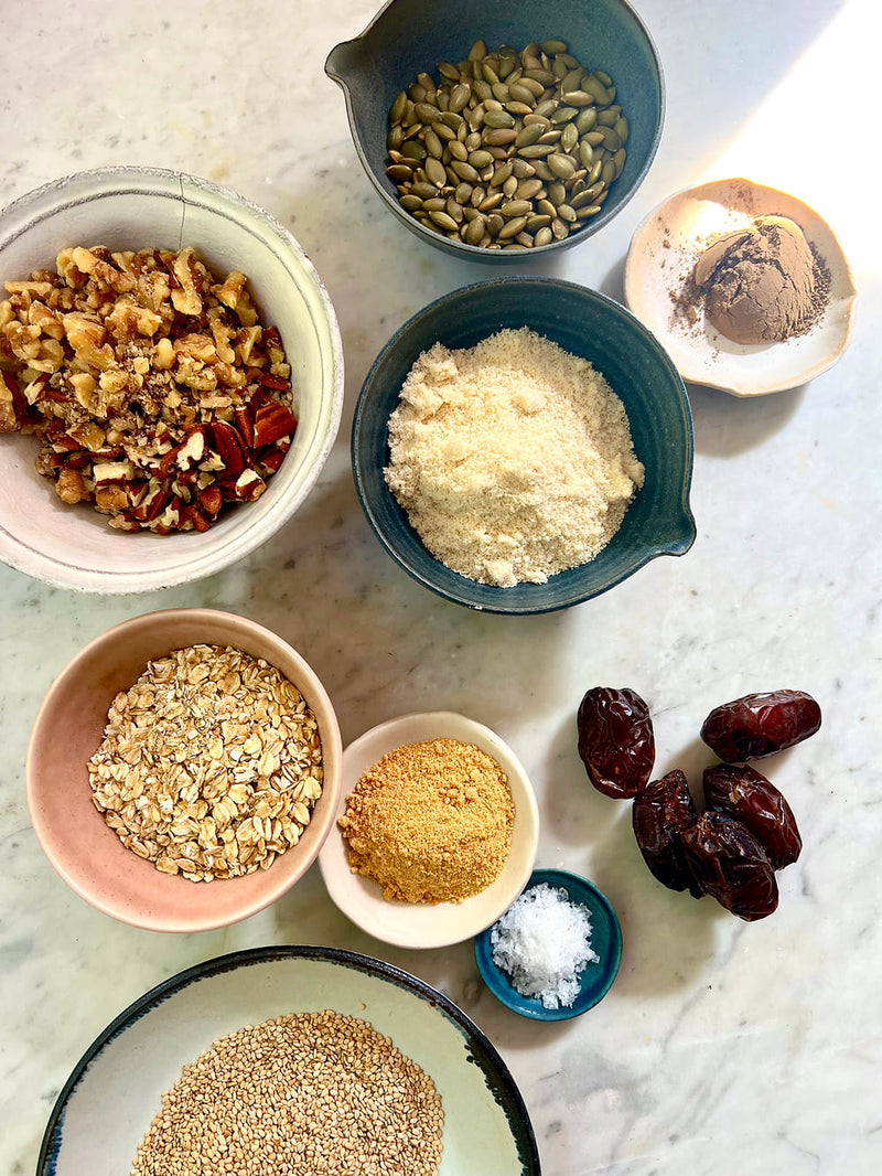 An overhead flat-lay of various ingredients in bowls on a light marble surface, including chopped nuts, a pale powder, seeds, dates, oats, and spices.