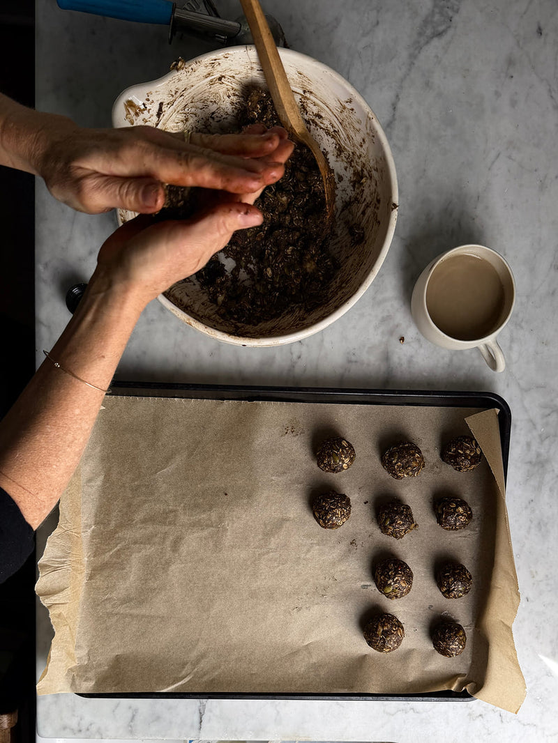 Top-down view of hands shaping cookie dough balls from a mixture in a white bowl, with a baking sheet of formed dough balls and a mug on a light marble surface.