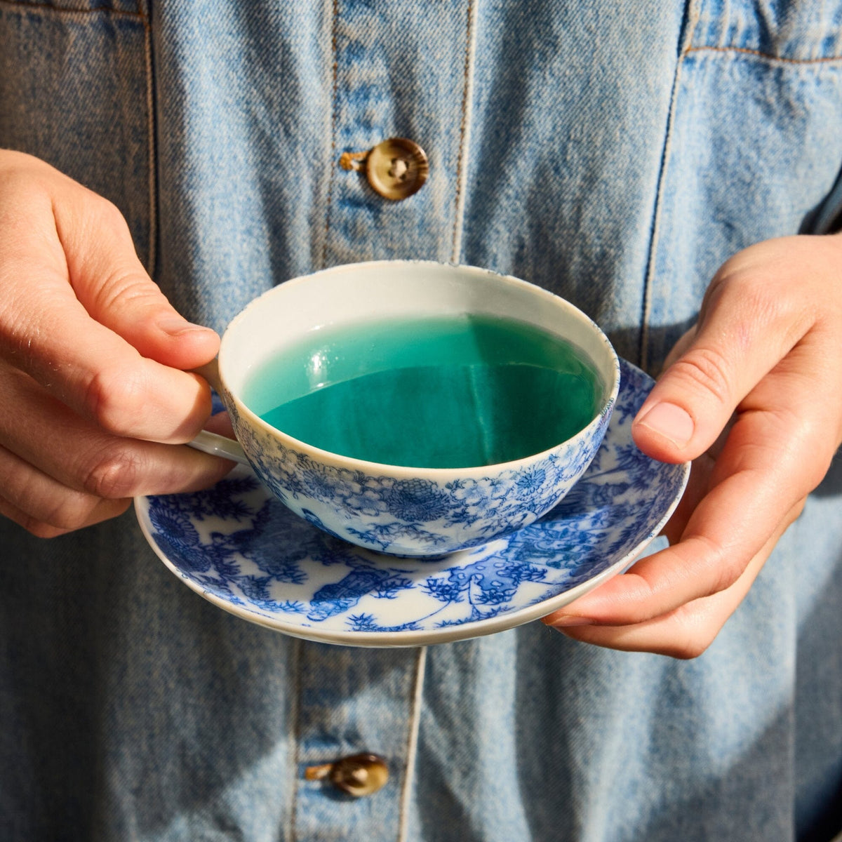 A person in a denim shirt holds a blue-and-white teacup and saucer filled with teal-colored liquid, likely Butterfly Pea Flower tea from Rishi Tea & Botanicals.