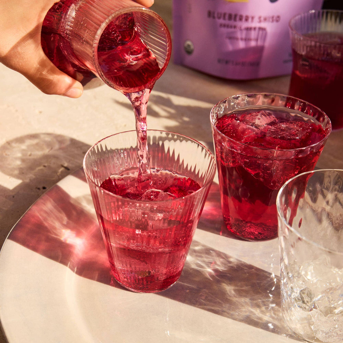 A hand pours a pink-purple fruity iced tea from a pitcher into a clear glass on a white plate, with other glasses of the same refreshing Rishi Tea & Botanicals drink nearby.