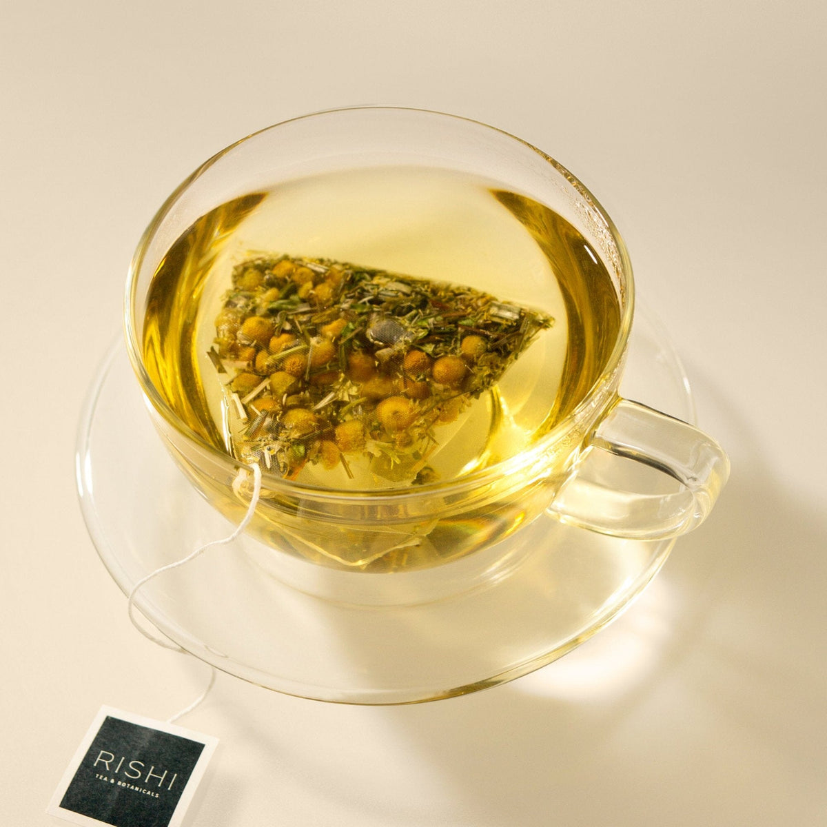 A clear glass cup of Chamomile Medley herbal tea from Rishi Tea & Botanicals, with a floral aroma, steeps inside and sits on a matching saucer. A "RISHI" tea tag is visible, inviting you to relax and unwind.
