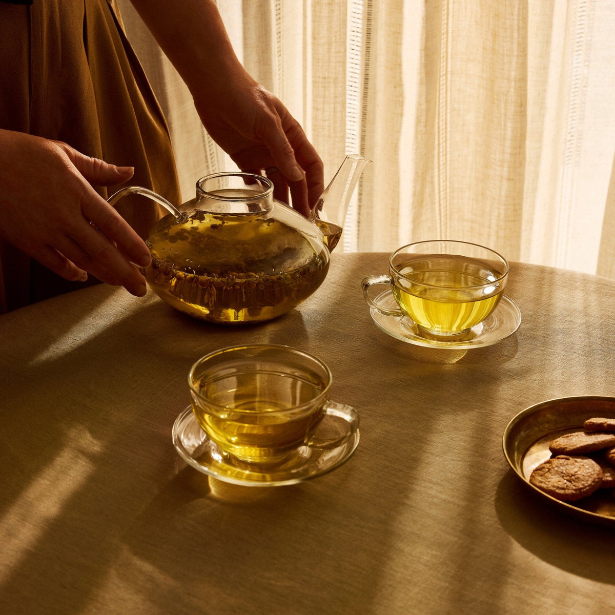 A person pours Rishi Tea & Botanicals Chamomile Medley from a glass teapot into clear cups on a round table with cookies, bathed in warm sunlight—a perfect, relaxing moment to enjoy the gentle floral aroma.