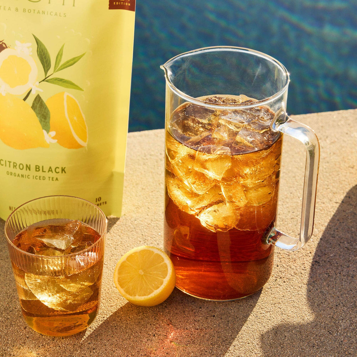 A glass pitcher and a glass of iced tea with ice cubes rest on a sunlit surface beside a halved lemon and Rishi Tea & Botanicals’ Citron Black Yunnan Dianhong black tea package. A shimmering pool appears in the background.
