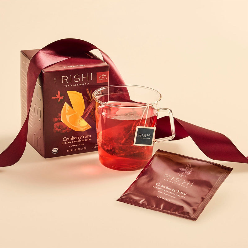 A glass mug of cranberry yuzu tea, a Rishi Tea & Botanicals box, and a sachet rest on a beige surface with a dark red ribbon, showcasing hints of hibiscus for a vibrant botanical accent.