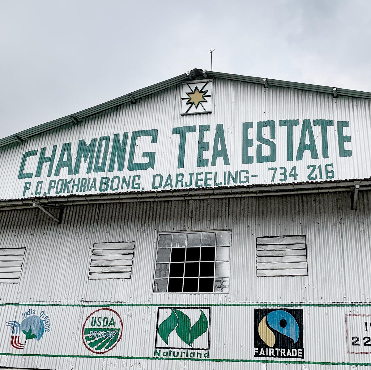 An industrial building with the sign "Rishi Tea & Botanicals" and various certification logos, including USDA Organic and Fairtrade. The building is in Darjeeling, as indicated by the address on the sign. Darjeeling First Flush Chamong EX1 is known for its notes of stone fruits and fragrant blossoms.