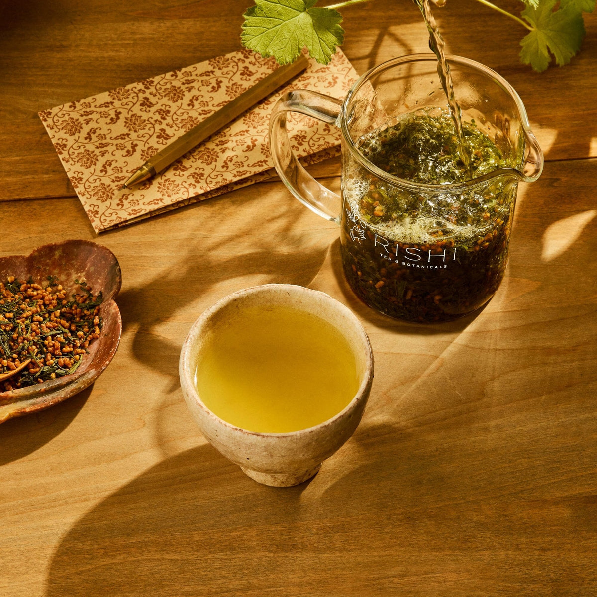 A cup of green tea from Rishi Tea & Botanicals rests on a wooden table beside a glass teapot of Genmaicha, a bowl of roasted sticky rice and tea leaves, and a patterned cloth.