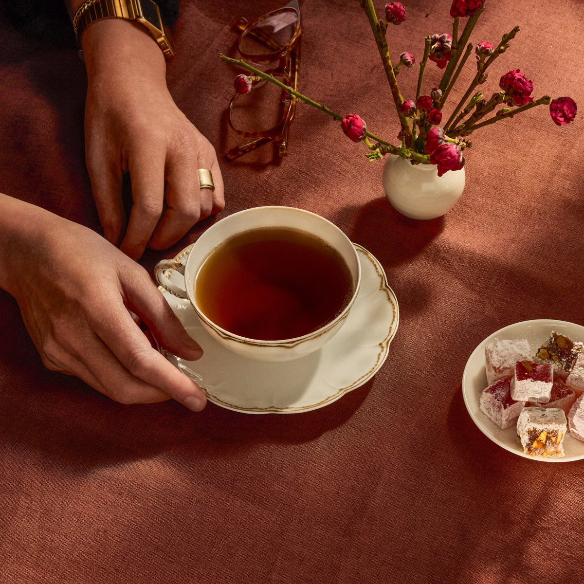 A person holds a cup of Rishi Tea & Botanicals Golden Yunnan tea on a saucer beside a small vase with pink flowers, eyeglasses, and a plate of Turkish delight on a reddish-brown tablecloth.