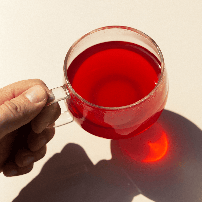 A hand holds a clear glass cup filled with bright red hibiscus liquid from Rishi Tea & Botanicals, casting a vibrant red shadow on a light surface.