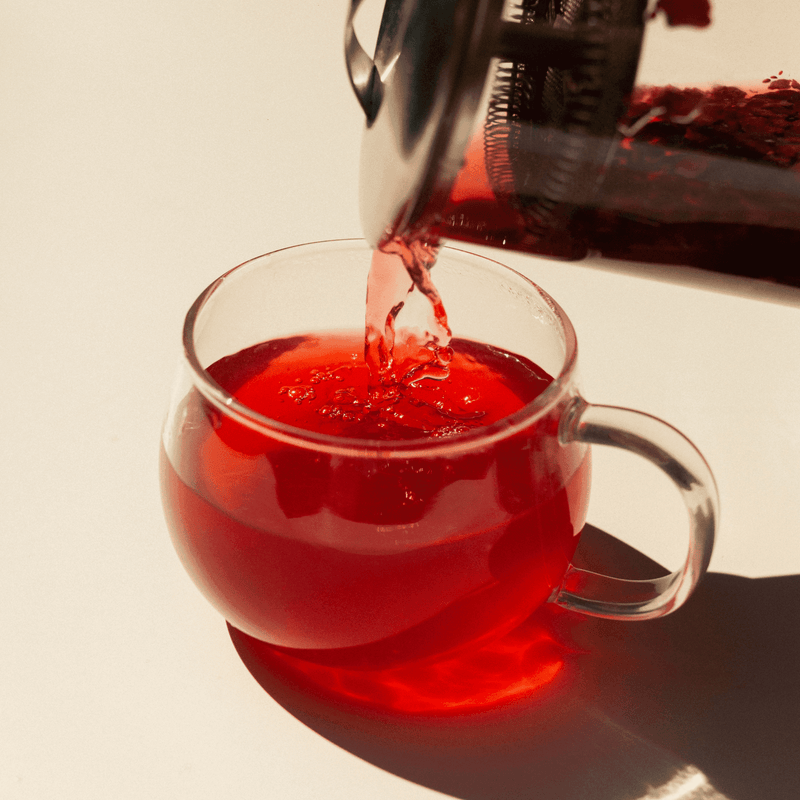 Rishi Tea & Botanicals' red herbal tea with schisandra berry is being poured from a glass French press into a clear teacup on a light surface.