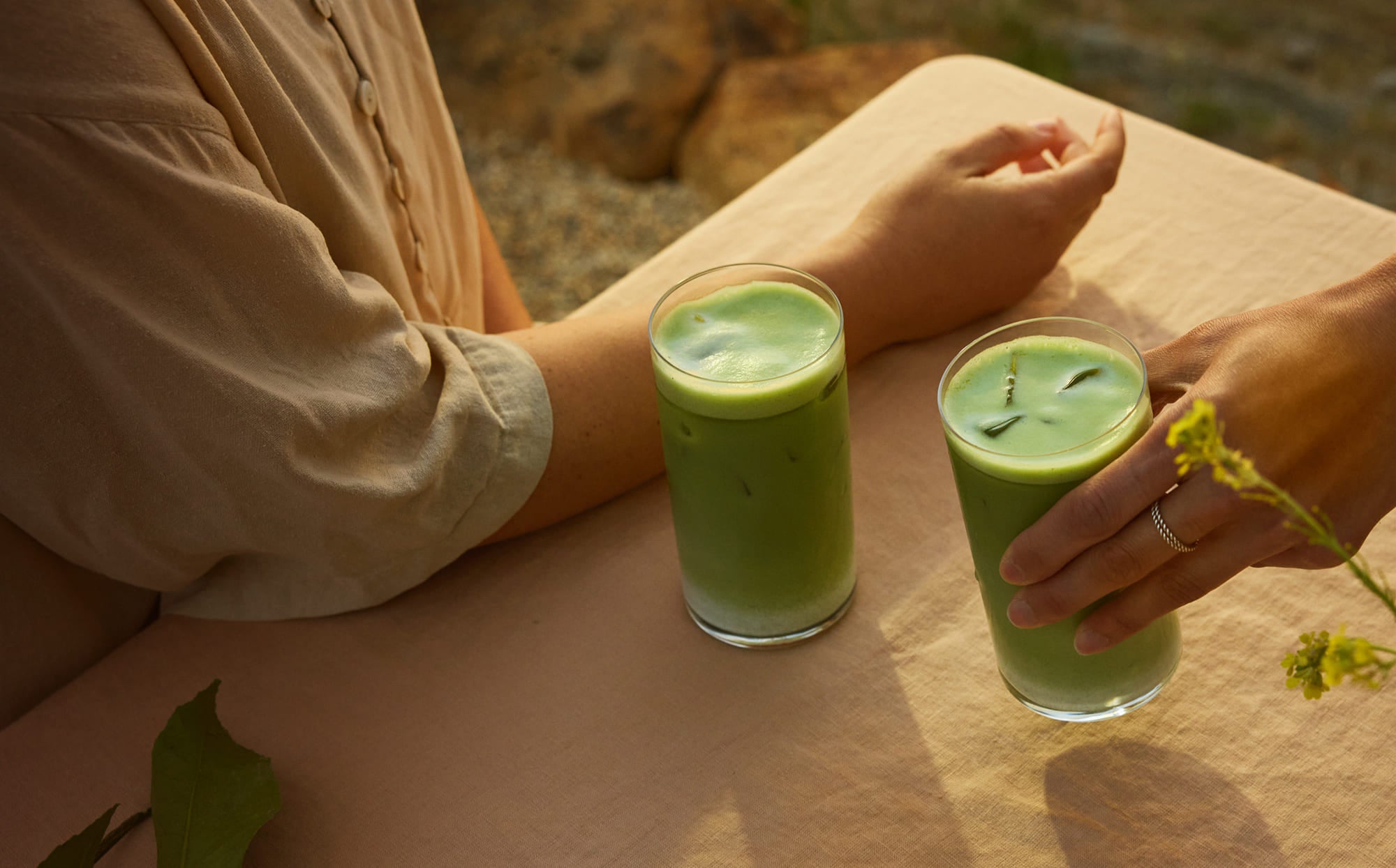 Two clear glasses filled with a bright green beverage sit on a beige tablecloth outdoors, with a person in a beige top seated at the table and another hand reaching toward the glass.
