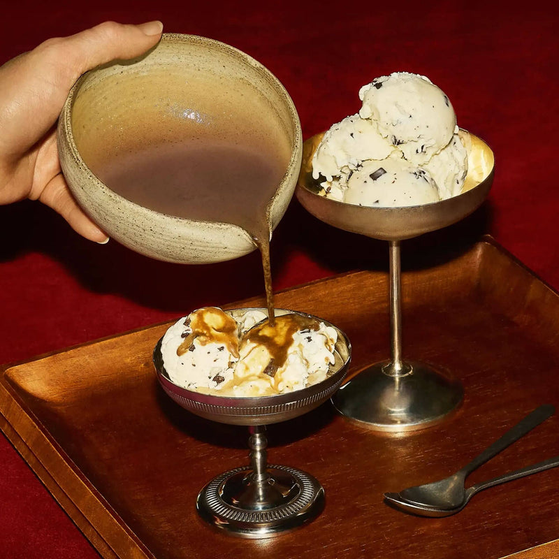Hand pouring roasted Houjicha sauce over ice cream in a silver dish, beside another dish, both on a wooden tray with two spoons, set against a red background.