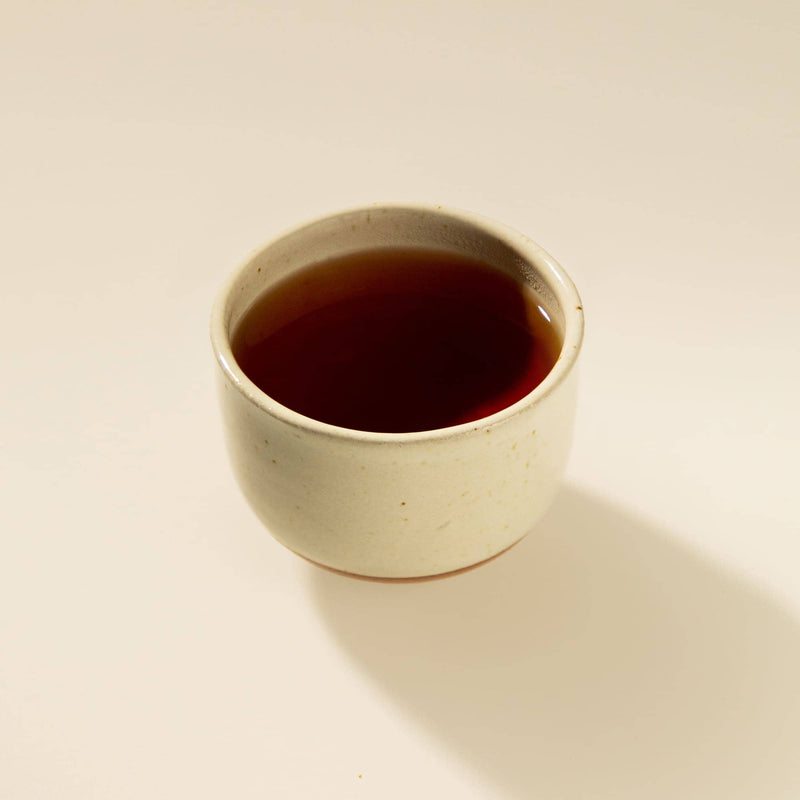 A small handmade ceramic cup filled with dark red tea, set against a plain off-white background with a soft shadow.