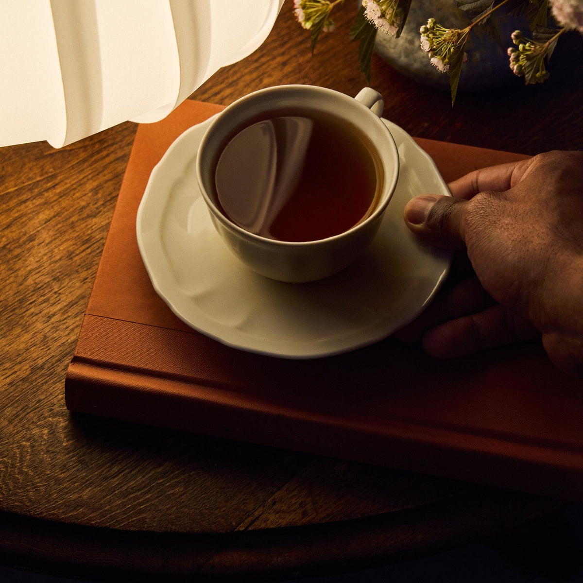 A hand holds a white cup of Rishi Tea & Botanicals' calming lavender tea on a saucer, resting atop a closed brown book on a wooden table beside a lamp and a vase of flowers.