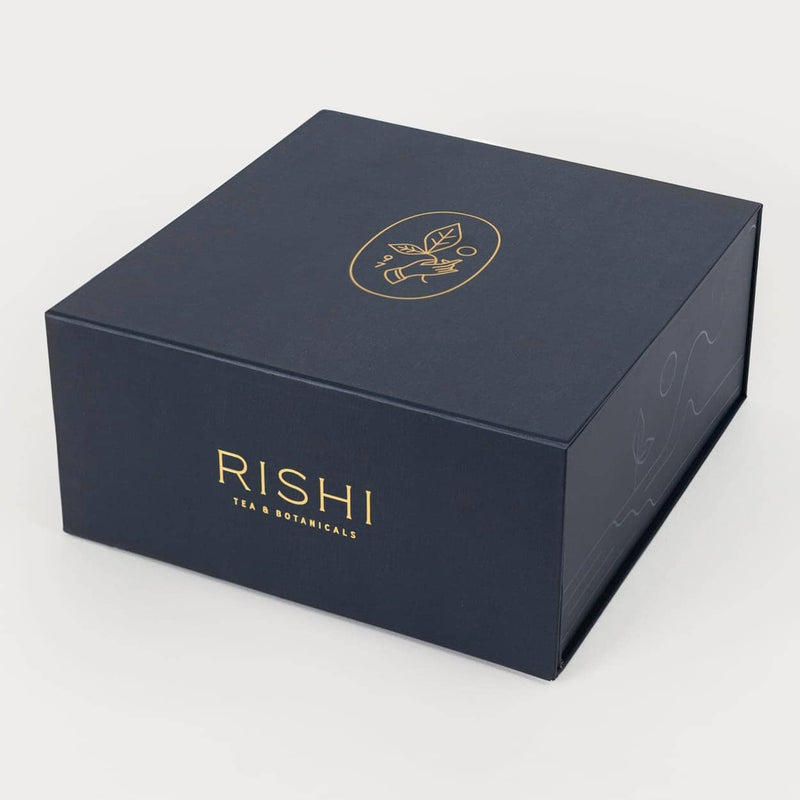A dark blue rectangular tea gift box with “RISHI TEA & BOTANICALS” printed in gold lettering on the front and a gold emblem featuring leaves and a teacup on the lid, perfect for presenting loose leaf tea or pairing with a glass teapot.