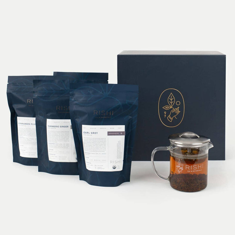 Three blue Rishi tea bags, a glass teapot with brewed loose leaf tea, and a large blue Rishi branded tea gift box are displayed on a white surface.