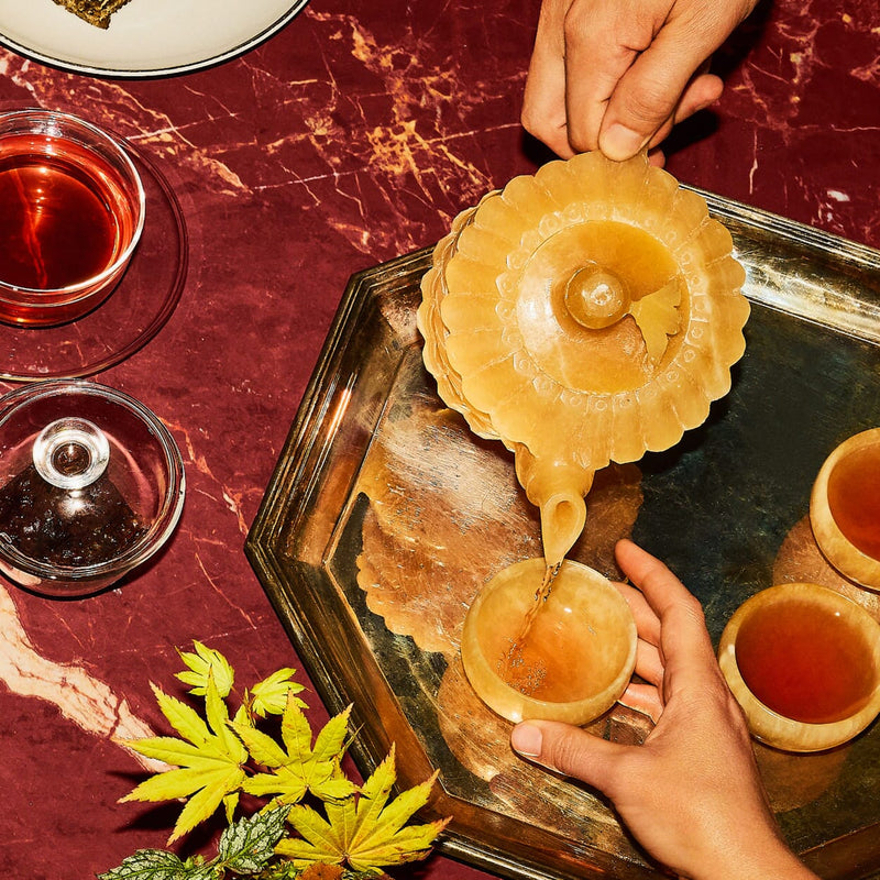 A person pours tea from a teapot into a cup on a metal tray with other filled cups, a green leaf, and glassware on a maroon marble surface.