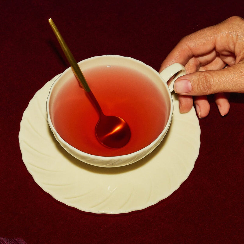 A hand holds a white teacup filled with red tea on a matching saucer. A gold spoon rests inside the cup, and the background is a dark red surface.
