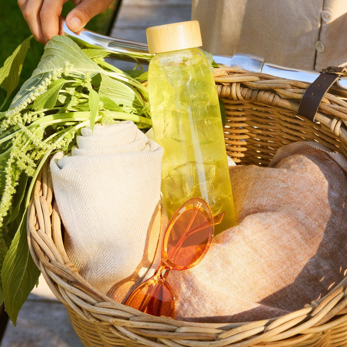 A woven basket holds a green leafy plant, a rolled towel, orange sunglasses, and a yellow Rishi Tea & Botanicals matcha drink bottle with a wooden lid.