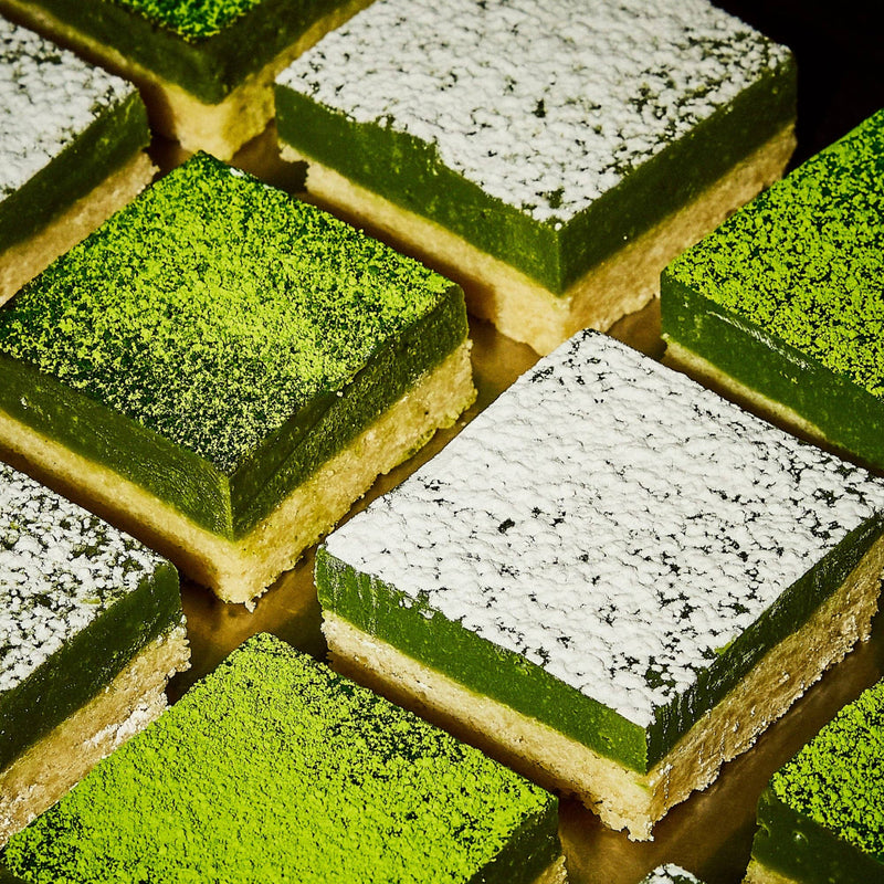 Rows of green matcha dessert bars with a yellow base, topped with either powdered sugar or matcha powder, arranged in a grid pattern.