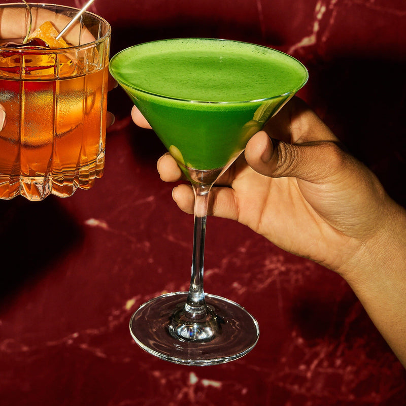 Two hands holding cocktails, one with an amber drink in a short glass, the other with a bright green drink in a martini glass, against a red marble background.