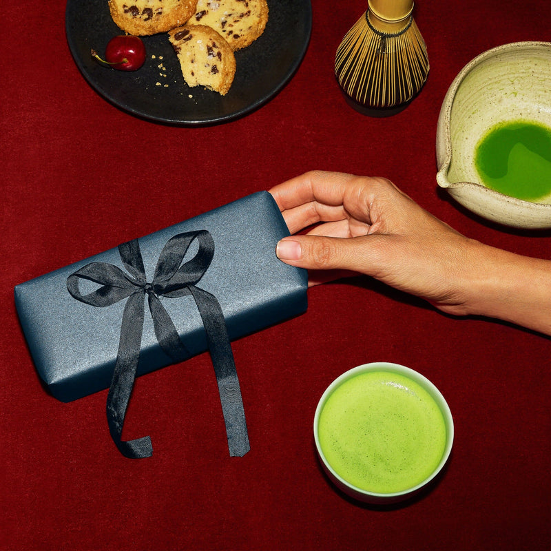 A hand holds a navy blue gift box with a black ribbon beside a cup of green tea, a bowl, cookies, a cherry, and a bamboo whisk on a red surface.