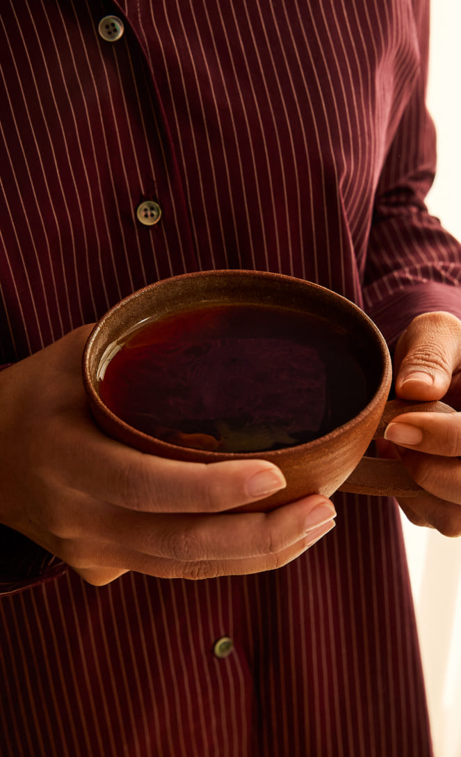 A close-up of a person wearing a burgundy button-up shirt with thin white vertical stripes, holding a small brown ceramic bowl containing a dark red liquid and a silver foil packet.