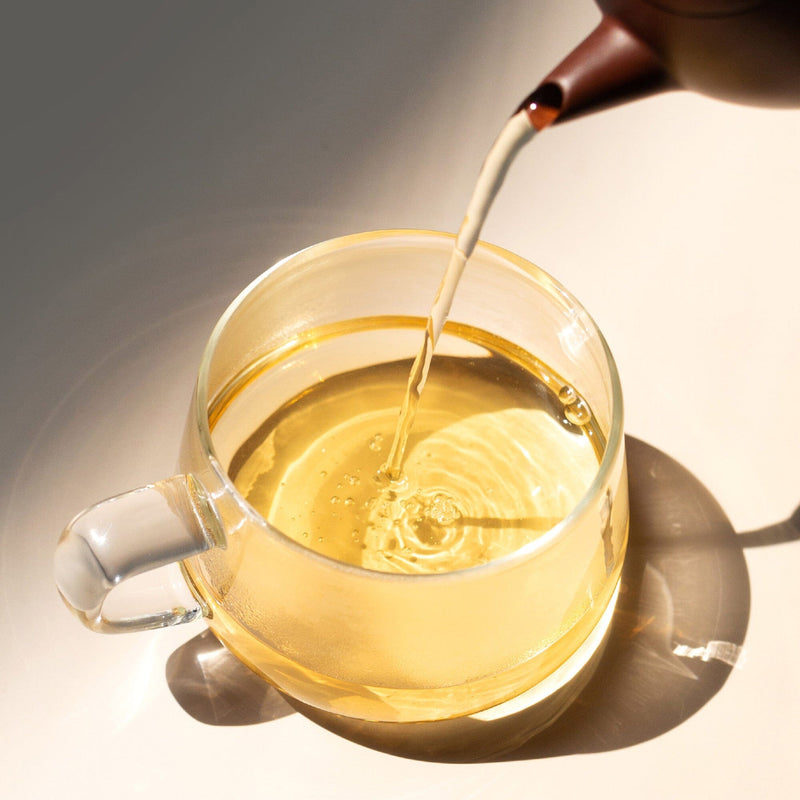 Clear tea with subtle Wuyi oolong notes from Rishi Tea & Botanicals is poured from a teapot into a transparent glass cup on a light surface, sunlight casting reflections and hints of orchid fragrance.