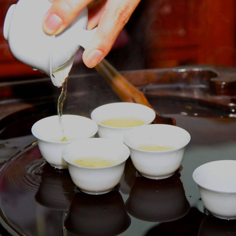 A hand pours creamy roast tea from a white teapot into small white teacups arranged in a circle on a reflective dark surface, creating a luxurious drinking experience.