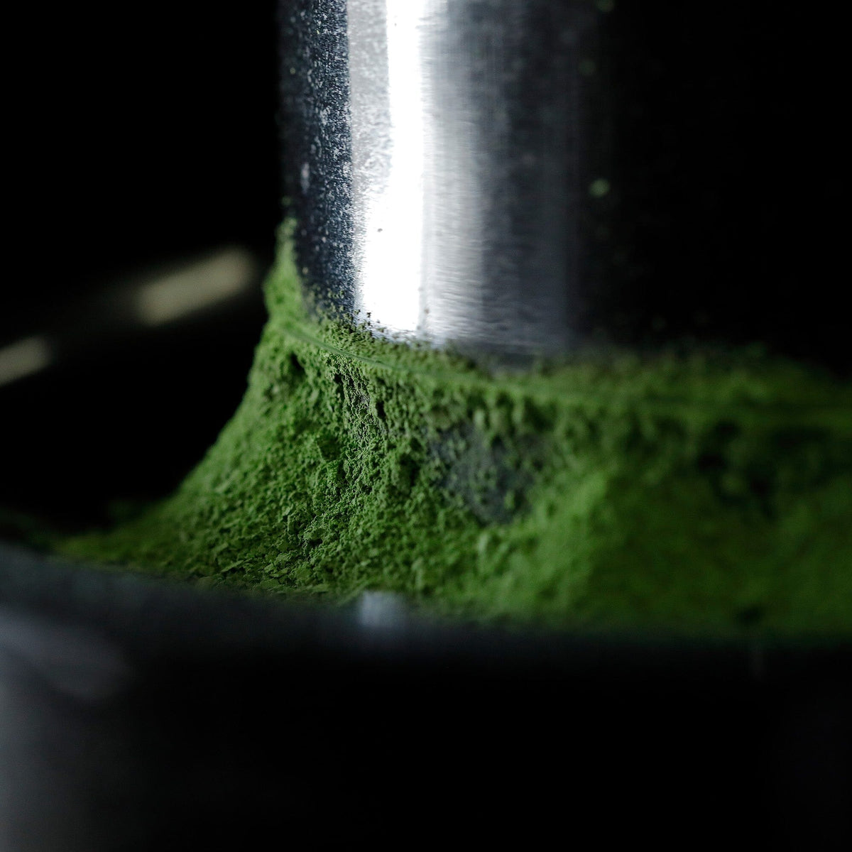 Close-up of green powder—likely matcha from Rishi Tea & Botanicals—gathering around a metallic object on a dark background, evoking the refined flavors championed by Hisaki Horii.