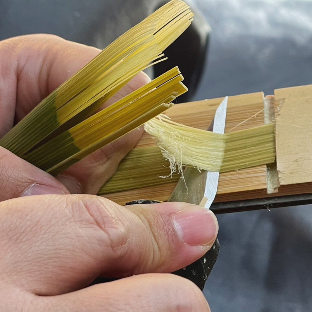 Someone uses a small knife to shave thin strips of green-yellow plant material held in a wooden clamp, possibly bamboo, perhaps crafting a Rishi Tea & Botanicals chasen for preparing Japanese tea or matcha.