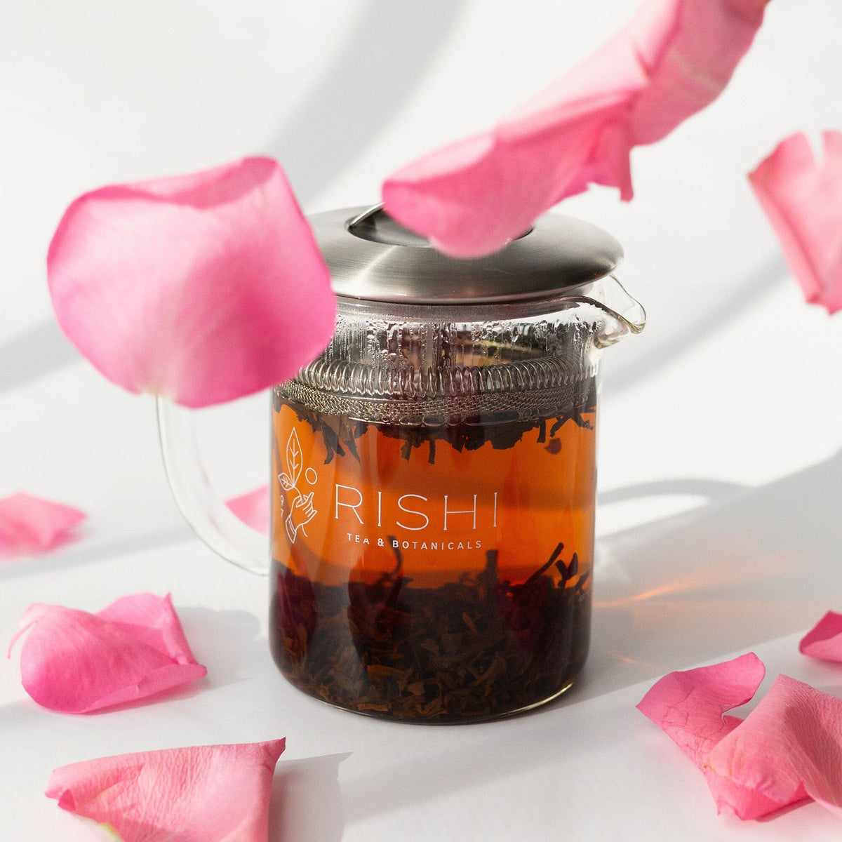 A glass teapot labeled "Rishi Tea & Botanicals" filled with Organic Dian Hong black tea rests on a white surface among pink rose petals, showcasing the brand's signature rose-scenting craftsmanship.