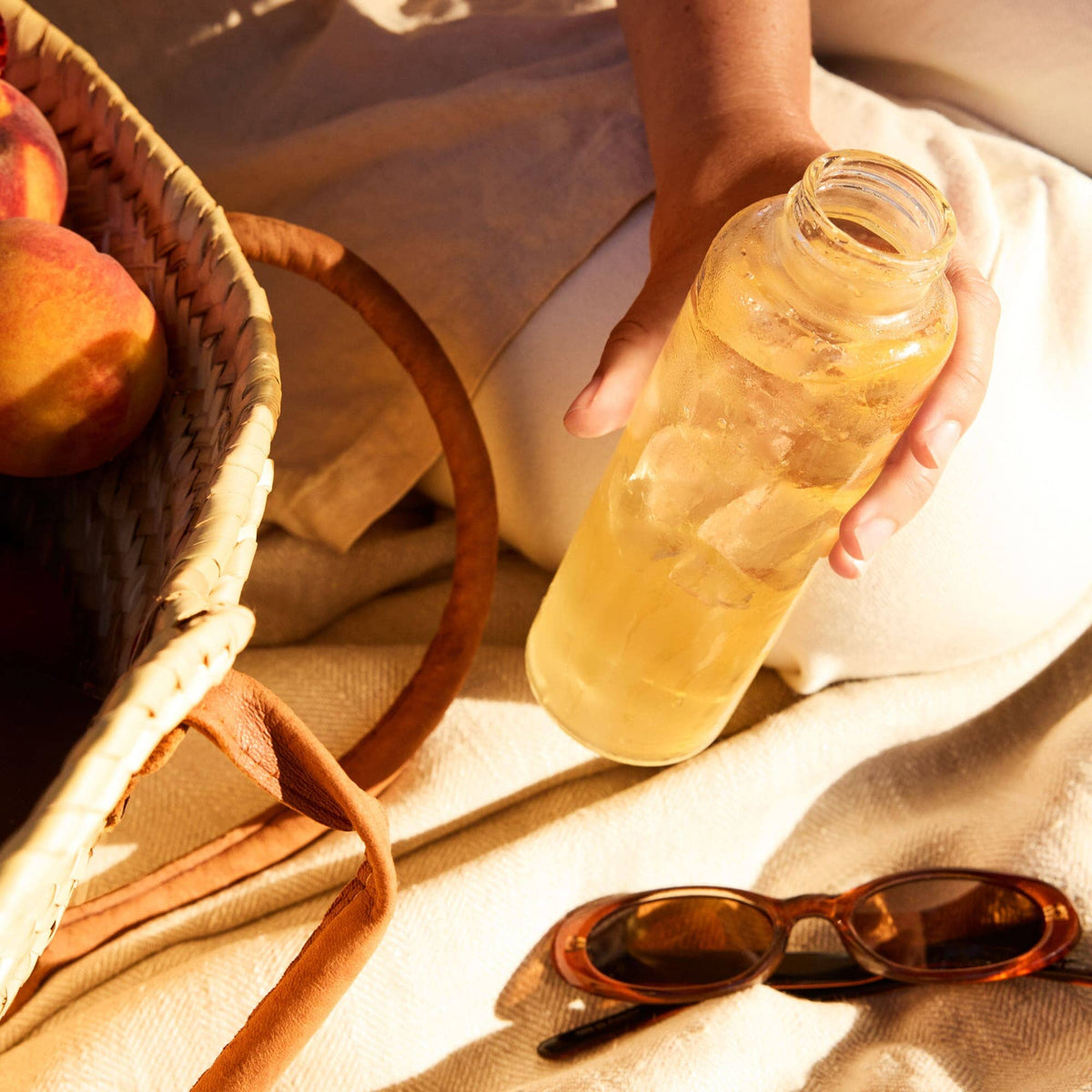 Person holding a bottle of iced tea next to a basket of peaches and sunglasses on a light fabric surface.