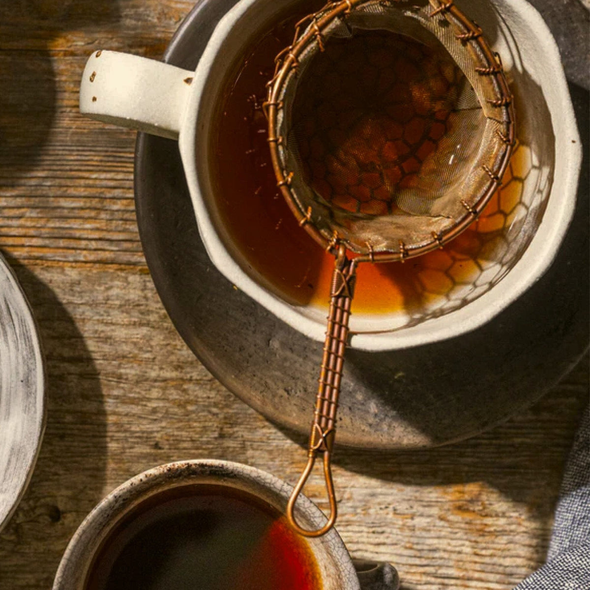 A metal tea strainer rests on a cup filled with dark Shu Pu'er tea from Rishi Tea & Botanicals, its earthy sweetness and hints of cocoa perfuming the air, placed on a wooden surface next to another cup.