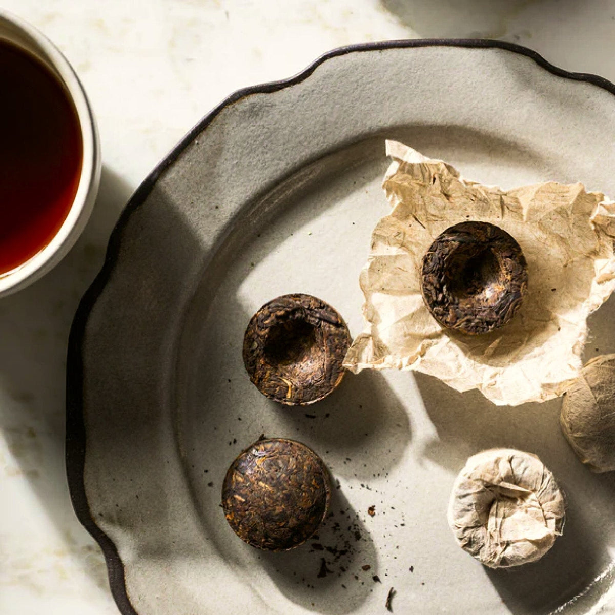 A cup of tea sits beside a plate showcasing round compressed tea leaves—some wrapped in paper, others unwrapped—on a light surface, making it perfect for savoring the exquisite vintage flavors of Rishi Tea & Botanicals’ traditional Pu'er tea.