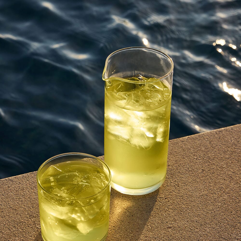 A glass and a pitcher of iced Sencha by Rishi Tea & Botanicals, an organic Japanese tea, rest on a sunlit concrete surface beside deep blue water.