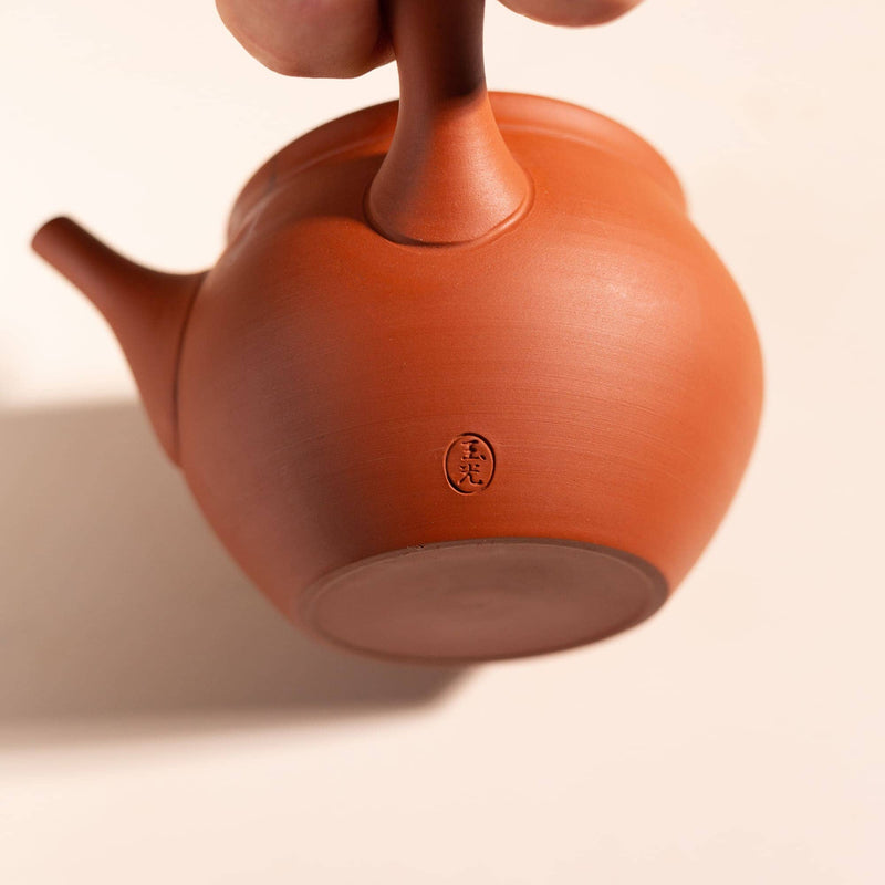 A close-up of a traditional clay teapot in a warm terracotta or reddish-brown color, held by a hand at the top. The teapot features a small circular embossed seal on the bottom center.