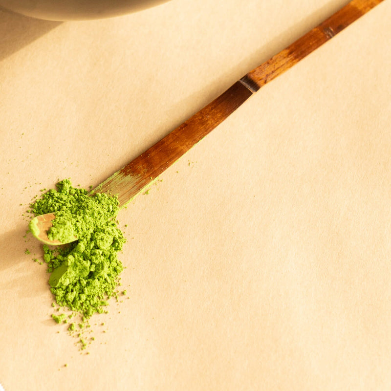 A Chashaku Bamboo Matcha Scoop from Rishi Tea & Botanicals with a pile of green matcha powder on a beige surface.