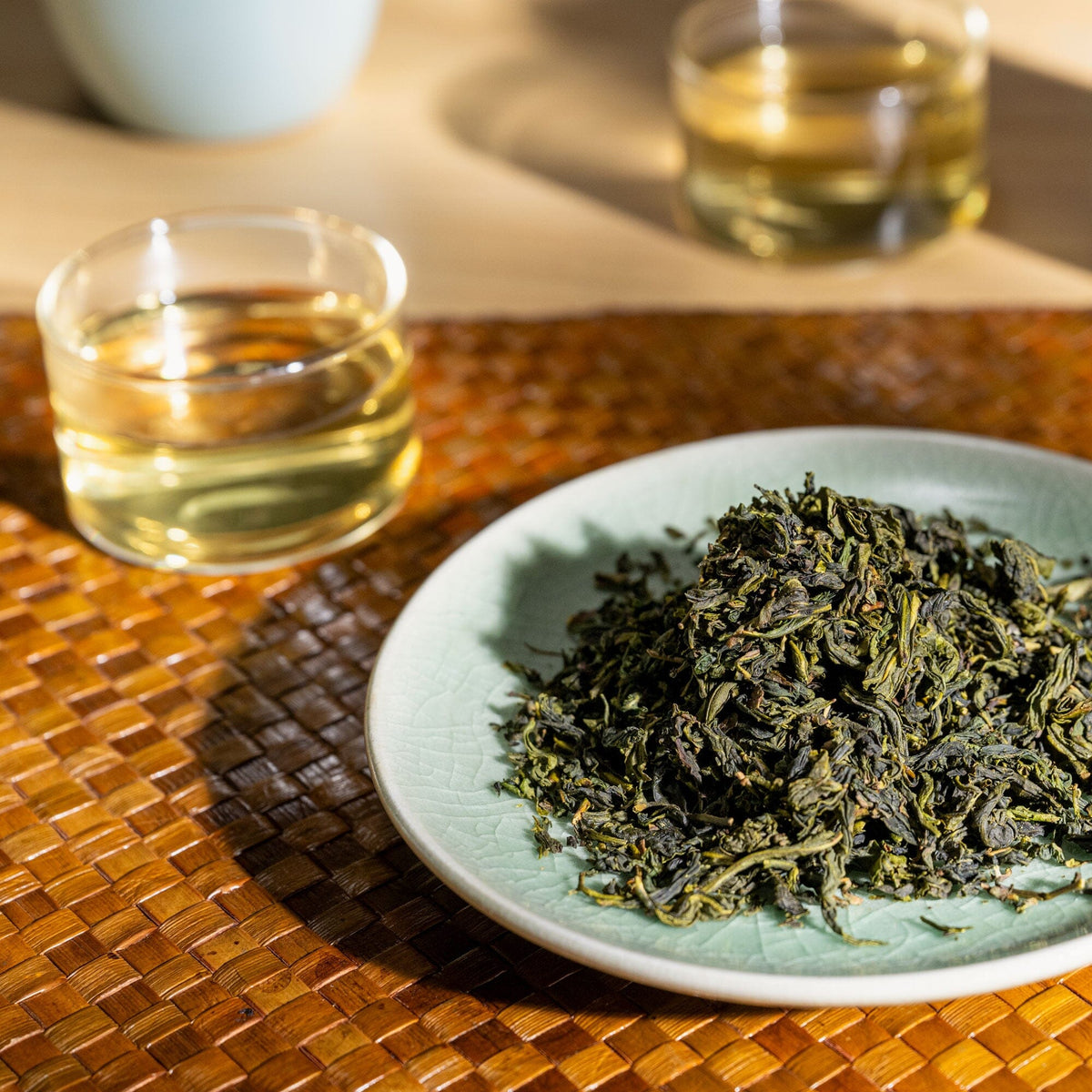 A plate of dried Bergamot Oolong leaves from Rishi Tea & Botanicals is on a woven mat next to two glasses of brewed oolong tea, with another cup in the background.