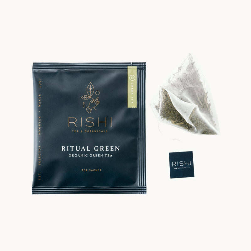 A sealed packet labeled “Rishi Ritual Green Organic Green Tea” lies next to a pyramid-shaped tea sachet with a tag reading “Rishi,” featuring premium green tea leaves sourced from the lush highlands of Northern Thailand and Myanmar.