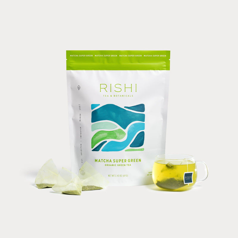 A bag of Rishi Tea & Botanicals Matcha Super Green sits beside two organic sachets and a glass mug of brewed green tea, featuring vibrant Japanese Sencha sourced from Kyushu Island.