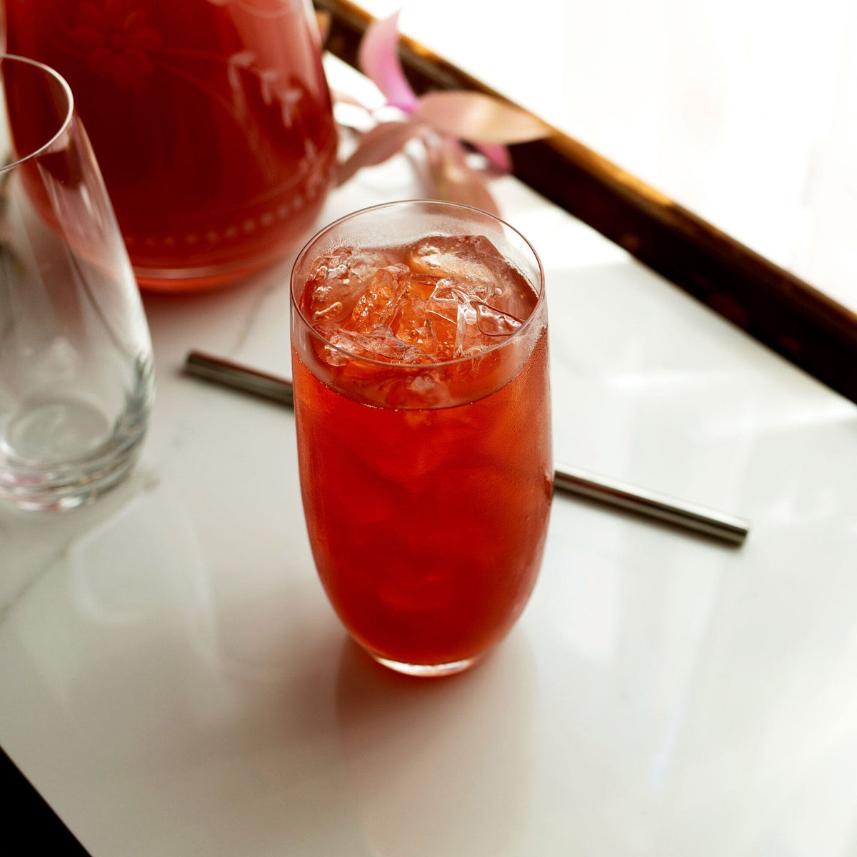 A glass of iced Passionfruit Hibiscus beverage made from Rishi Tea & Botanicals powder sits on a white surface beside an empty glass and a steel straw in front of a sunlit window.